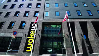 Exterior of a modern, multi-story hotel with dark brick facade and numerous windows. Two red and white flags are displayed in front. A sign with wakeup is prominent on the building, near a large glass entrance.