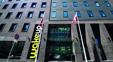 Exterior of a modern, multi-story hotel with dark brick facade and numerous windows. Two red and white flags are displayed in front. A sign with wakeup is prominent on the building, near a large glass entrance.