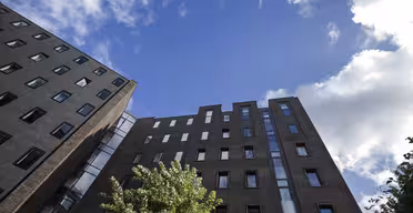 Upward view of two modern, dark-colored apartment buildings against a clear blue sky with scattered clouds. A green tree is partially visible in the foreground.