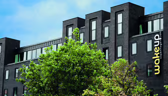 A modern black building with multiple windows, partially obscured by green foliage. The building has wakeup copenhagen written vertically in yellow and white on the right side. The sky is blue with scattered clouds.