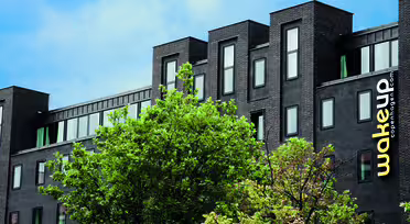 A modern black building with multiple windows, partially obscured by green foliage. The building has wakeup copenhagen written vertically in yellow and white on the right side. The sky is blue with scattered clouds.