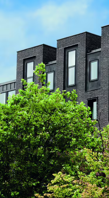 A modern black building with multiple windows, partially obscured by green foliage. The building has wakeup copenhagen written vertically in yellow and white on the right side. The sky is blue with scattered clouds.
