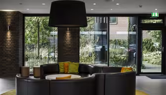 A modern lobby features a semi-circular black leather sofa with green and orange cushions. Large windows reveal lush green foliage outside. A large black lamp hangs overhead, and brick walls add texture to the space.