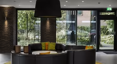 A modern lobby features a semi-circular black leather sofa with green and orange cushions. Large windows reveal lush green foliage outside. A large black lamp hangs overhead, and brick walls add texture to the space.
