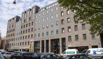 Street view of a modern brick and concrete building with large windows. Vehicles, including a van and several cars, are parked along the street. A tree branches over from the right, and the sky is partly cloudy.
