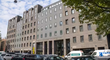 Street view of a modern brick and concrete building with large windows. Vehicles, including a van and several cars, are parked along the street. A tree branches over from the right, and the sky is partly cloudy.