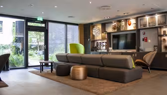 Modern lounge with brown modular couches, circular leather stools, and a large TV. Walls feature art and shelves with decor. A glass door opens to a garden, letting in natural light. Neutral tones and a cozy rug enhance the inviting atmosphere.