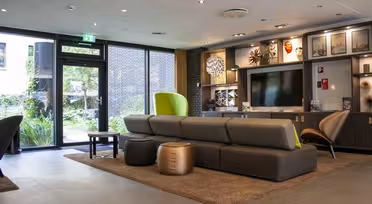 Modern lounge with brown modular couches, circular leather stools, and a large TV. Walls feature art and shelves with decor. A glass door opens to a garden, letting in natural light. Neutral tones and a cozy rug enhance the inviting atmosphere.