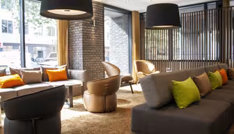 Modern hotel lobby with gray sofas and brown chairs. Bright green and orange pillows add a pop of color. Large black pendant lights hang from the ceiling. Sunlight streams through floor-to-ceiling windows, illuminating the brick and wood accents.
