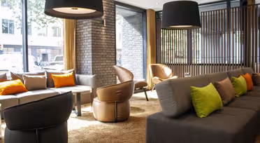 Modern hotel lobby with gray sofas and brown chairs. Bright green and orange pillows add a pop of color. Large black pendant lights hang from the ceiling. Sunlight streams through floor-to-ceiling windows, illuminating the brick and wood accents.