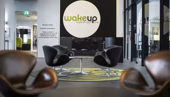 Modern hotel lobby with sleek brown chairs, circular tables, and a large logo on the wall that reads Wakeup Copenhagen. Glass walls allow natural light to fill the space, highlighting the contemporary decor.