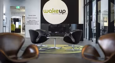 Modern hotel lobby with sleek brown chairs, circular tables, and a large logo on the wall that reads Wakeup Copenhagen. Glass walls allow natural light to fill the space, highlighting the contemporary decor.