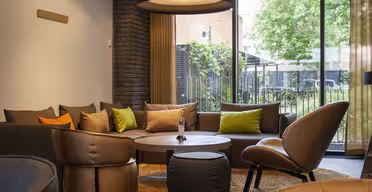 A modern living room with a large sofa with gray and lime green pillows, round wooden coffee table, and brown upholstered chairs. A large pendant light hangs above. The room has a brick wall and a window with a view of a garden outside.