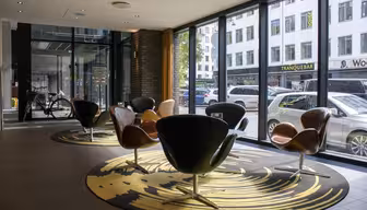 A modern lounge area with stylish, curved chairs on a patterned rug. Large windows reveal a busy city street outside. Various shops and parked cars are visible through the glass. The space is well-lit and contemporary.