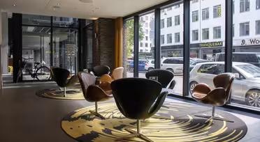 A modern lounge area with stylish, curved chairs on a patterned rug. Large windows reveal a busy city street outside. Various shops and parked cars are visible through the glass. The space is well-lit and contemporary.