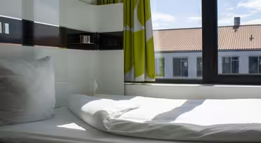 A minimalist bedroom with a neatly made single bed by a window. Sunlight streams in, illuminating the white bedding. The window offers a view of neighboring buildings and a bright sky. Green and white curtains add a touch of color.