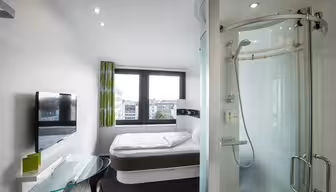 Modern hotel room with a bed by large windows, a glass shower enclosure, wall-mounted TV, small glass desk with a chair, and a view of buildings outside. Minimalist decor with white walls and a green curtain.
