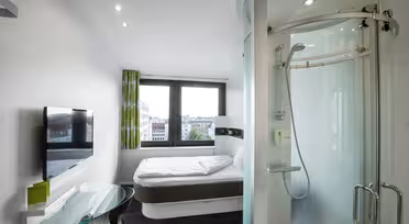 Modern hotel room with a bed by large windows, a glass shower enclosure, wall-mounted TV, small glass desk with a chair, and a view of buildings outside. Minimalist decor with white walls and a green curtain.