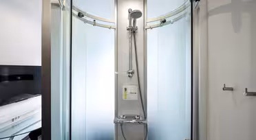 A modern, glass-enclosed shower booth with frosted sliding doors, a wall-mounted showerhead, and control panel. The sleek design features metallic handles and minimalistic fixtures, situated in a bright space with visible furniture in the background.