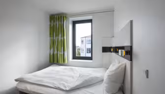 A small, minimalist bedroom features a neatly made bed with white linens and pillows against a white wall. A window with green and white patterned curtains lets in natural light, highlighting the simple yet modern decor.