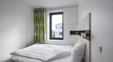 A small, minimalist bedroom features a neatly made bed with white linens and pillows against a white wall. A window with green and white patterned curtains lets in natural light, highlighting the simple yet modern decor.