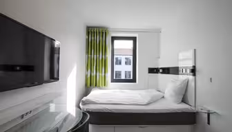 A small, modern bedroom with a single bed, white linens, and a green patterned curtain by the window. A wall-mounted flat-screen TV is opposite the bed, and a glass side table is beside it. The window shows a view of neighboring buildings.