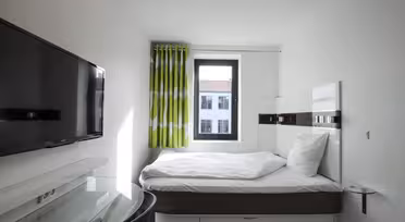 A small, modern bedroom with a single bed, white linens, and a green patterned curtain by the window. A wall-mounted flat-screen TV is opposite the bed, and a glass side table is beside it. The window shows a view of neighboring buildings.