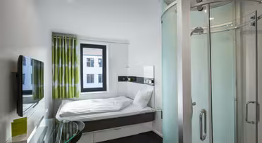 Modern hotel room with a single bed, white walls, and a round glass shower in the corner. A green and white curtain frames a window, and a wall-mounted TV is beside a glass desk with a green booklet.