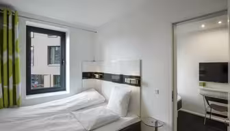 Modern hotel room with a bed, white linens, and green and white curtains. A window overlooks a building. Theres a small glass table beside the bed. An adjoining room with a TV and desk is visible through an open door.