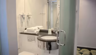 Modern bathroom with a round sink, wall-mounted hairdryers, and a glass shower enclosure. Towels are neatly stacked beside the sink. The floor features a circular pattern, and natural light brightens the space through frosted glass.