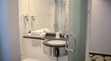 Modern bathroom with a round sink, wall-mounted hairdryers, and a glass shower enclosure. Towels are neatly stacked beside the sink. The floor features a circular pattern, and natural light brightens the space through frosted glass.