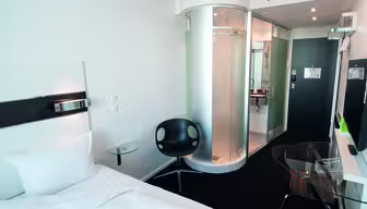 Modern hotel room with a white single bed, black circular chair, glass-topped desk, and wall-mounted TV. The room features a unique cylindrical glass shower near the entrance, and minimalist decor with white walls and contemporary furnishings.
