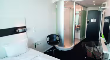 Modern hotel room with a white single bed, black circular chair, glass-topped desk, and wall-mounted TV. The room features a unique cylindrical glass shower near the entrance, and minimalist decor with white walls and contemporary furnishings.