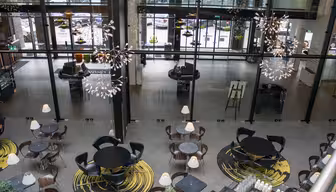 A modern indoor café with round tables and chairs arranged on circular rugs. Large glass windows provide a view of the street outside. Elegant chandeliers hang from the ceiling, and soft lighting creates a cozy atmosphere.