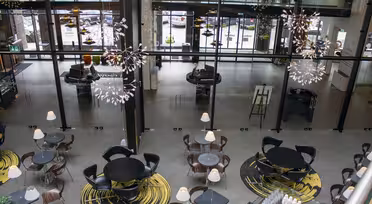 A modern indoor café with round tables and chairs arranged on circular rugs. Large glass windows provide a view of the street outside. Elegant chandeliers hang from the ceiling, and soft lighting creates a cozy atmosphere.