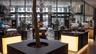 Modern cafe interior with a tree in the center and illuminated counters. Contemporary decor includes hanging star-shaped lights and plants. Glass walls provide a view of the outside.