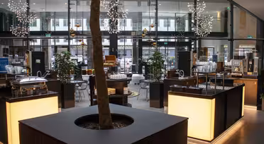 Modern cafe interior with a tree in the center and illuminated counters. Contemporary decor includes hanging star-shaped lights and plants. Glass walls provide a view of the outside.