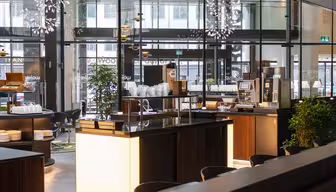 Modern, well-lit cafeteria interior with wooden finishes and glass walls. The counter displays plates and a coffee machine. Large windows reveal a view of the street outside, while plants add greenery to the spacious area.