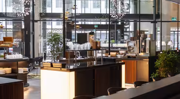 Modern, well-lit cafeteria interior with wooden finishes and glass walls. The counter displays plates and a coffee machine. Large windows reveal a view of the street outside, while plants add greenery to the spacious area.
