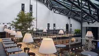 Modern, spacious indoor café with glass roof and metal beams. Features wooden floors, two levels, and tables with white lamps. A large indoor tree and framed artwork add to the decor. Natural light fills the space.