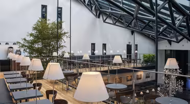 Modern, spacious indoor café with glass roof and metal beams. Features wooden floors, two levels, and tables with white lamps. A large indoor tree and framed artwork add to the decor. Natural light fills the space.