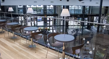 Modern indoor café with wooden floors and metal-framed tables and chairs. Each table has a small, lit lamp. Large glass windows overlook a street with parked cars. A railing separates the seating area from the view below.