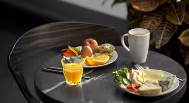 A small round table with a breakfast setting: a plate of fruit, a cup of coffee, a glass of orange juice, and a plate with bread, cheese, and vegetables. The background features a plant with variegated leaves.