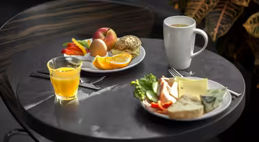 A round table with two breakfast plates; one includes an egg, orange slices, apple, and bread roll, the other has sliced vegetables, cheese, and a bread roll with butter. Accompanied by a cup of coffee and a glass of orange juice.
