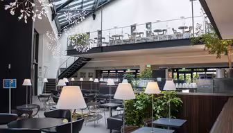 A modern, spacious restaurant interior with a high ceiling, featuring black tables and chairs. A mezzanine level with additional seating is visible. White pendant lights hang from the ceiling, and plants are placed throughout for decoration.