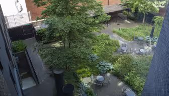 Aerial view of a courtyard with brick pathways, surrounded by lush greenery and trees. Several round metal tables with chairs are placed on the paths. A brick wall and buildings are in the background.
