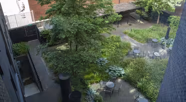 Aerial view of a courtyard with brick pathways, surrounded by lush greenery and trees. Several round metal tables with chairs are placed on the paths. A brick wall and buildings are in the background.