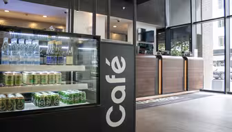 Modern café interior with a glass display filled with bottled water and canned drinks. A sleek wooden counter is behind it. Large windows let in natural light, revealing a view of greenery outside. The word Café is prominently displayed on the sign.