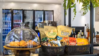 A display of snacks on a counter, including croissants under a glass dome, bags of chips, peanuts, and candy in baskets. Refrigerators with beverages are in the background, and green plants hang overhead.