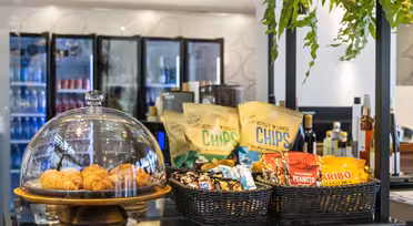 A display of snacks on a counter, including croissants under a glass dome, bags of chips, peanuts, and candy in baskets. Refrigerators with beverages are in the background, and green plants hang overhead.